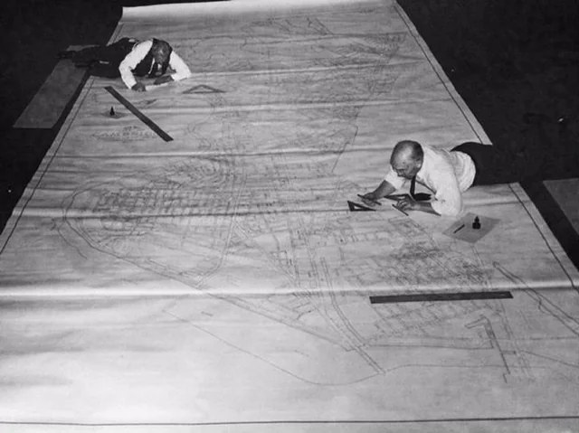 Two cartographers lying on a large map spread on the floor, with T-squares and ink.